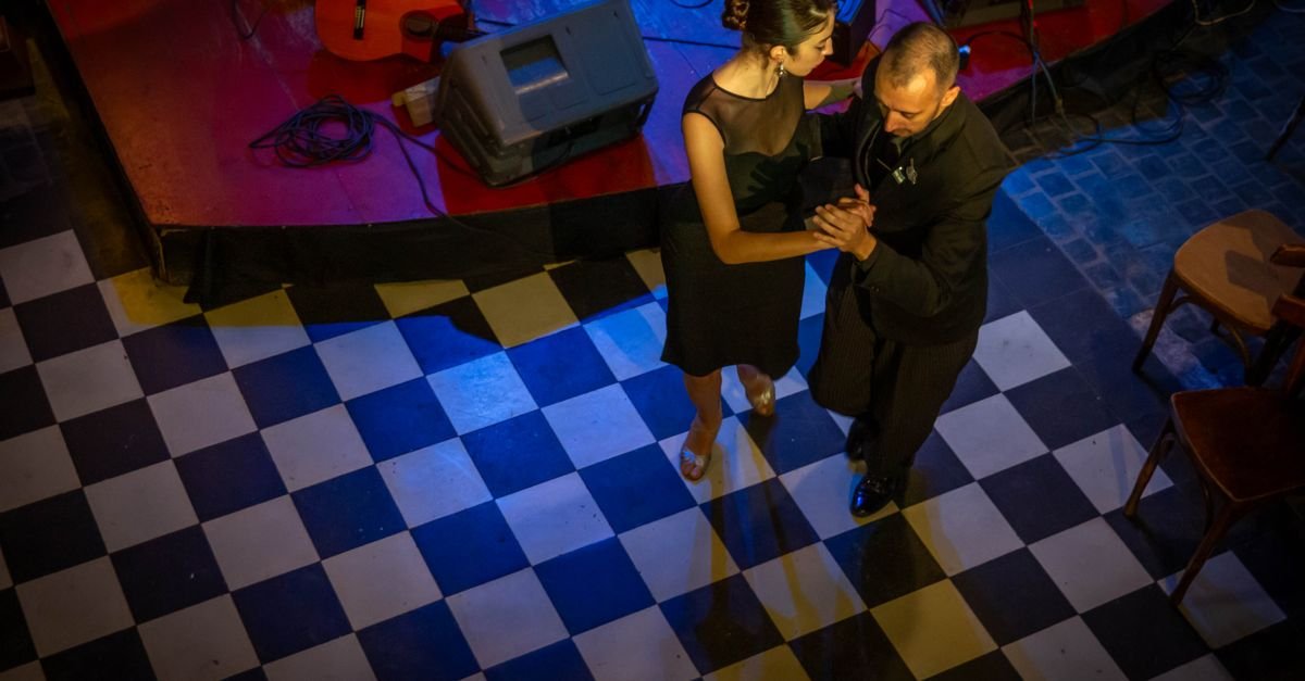 Casal dança tango em salão com piso quadriculado e iluminação azul