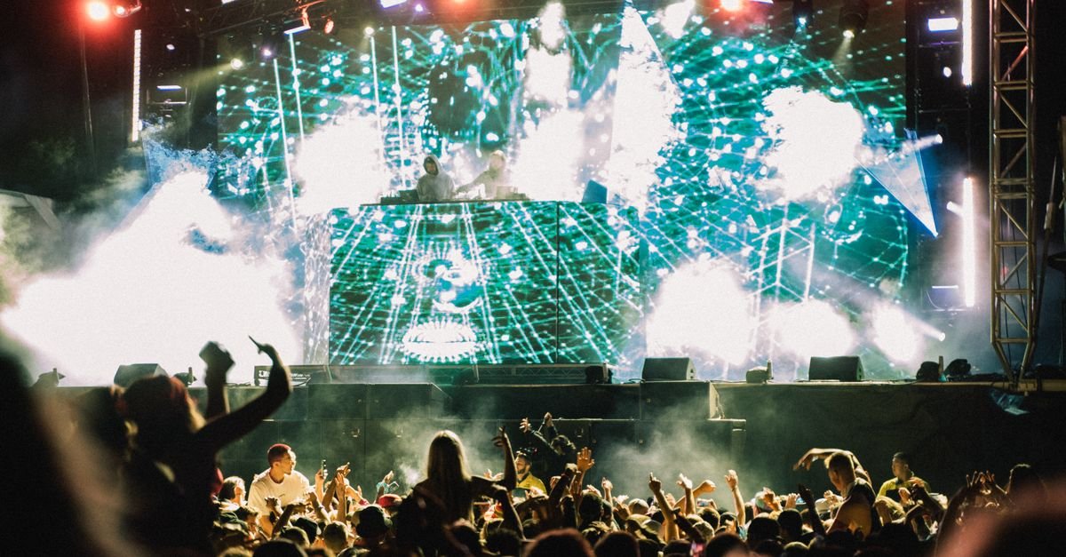 Público de festival de música eletrônica em frente a palco com painel de LED e efeitos de fumaça