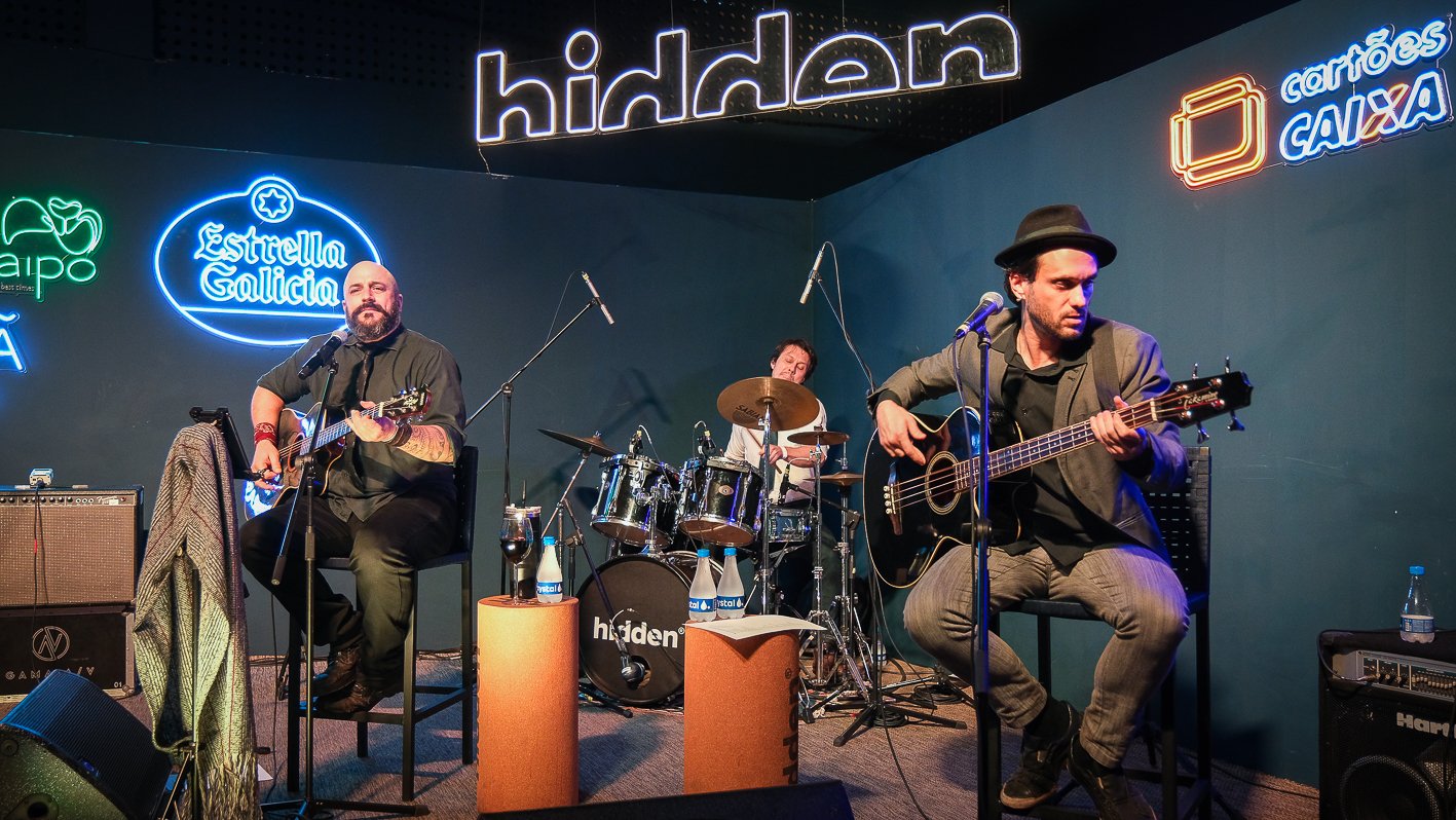 Tres musicos do Acustico Navaranda tocam violao, baixo e bateria no palco do Hidden Brasilia, sob o letreiro luminoso da casa