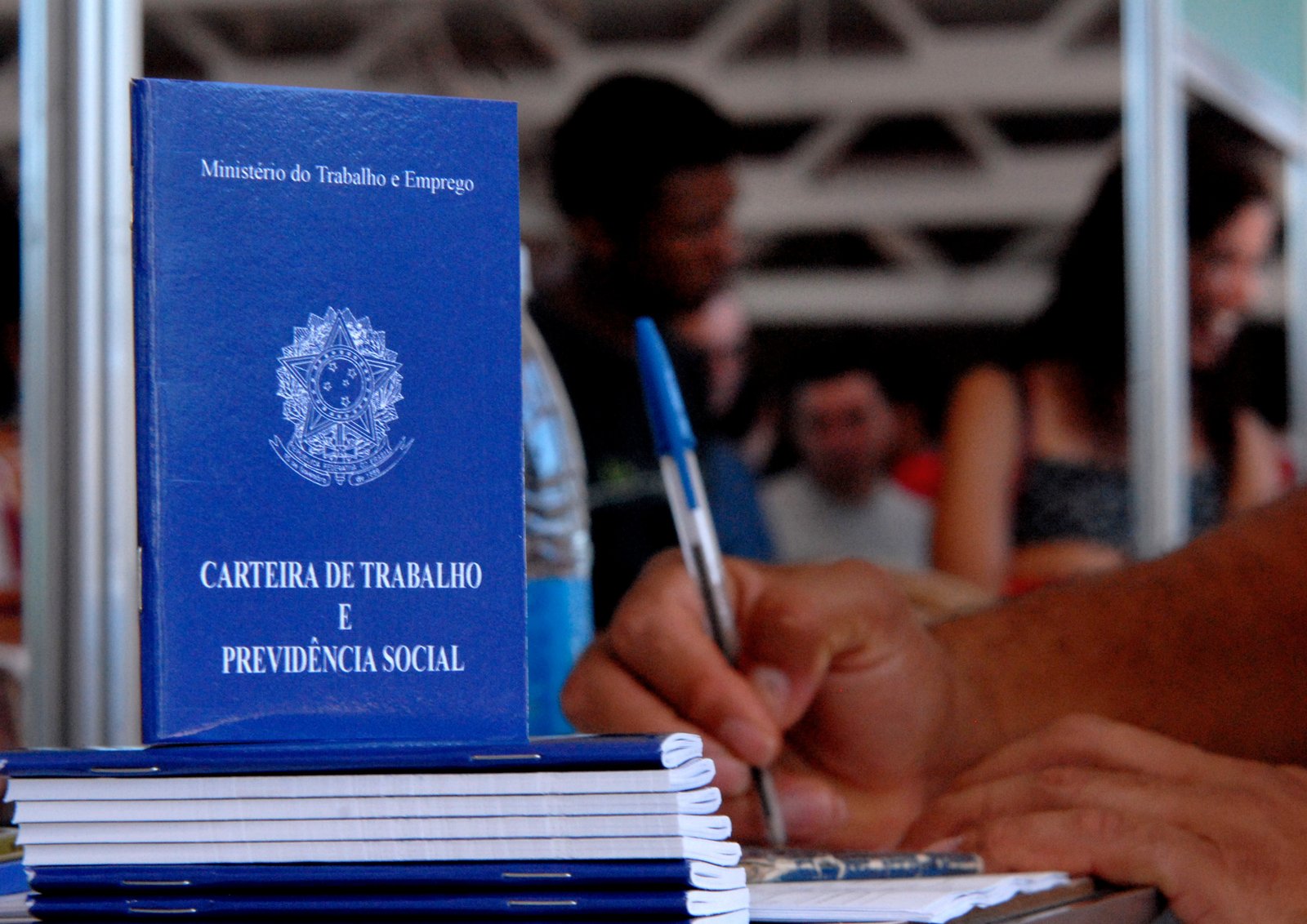 Carteiras de trabalho empilhadas sobre uma mesa de atendimento, com uma pessoa preenchendo um formulário
