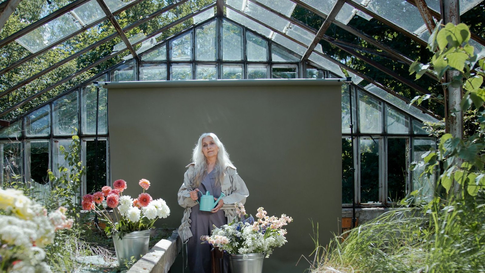 Mulher de cabelos grisalhos segura um regador entre vasos de flores dentro de uma estufa