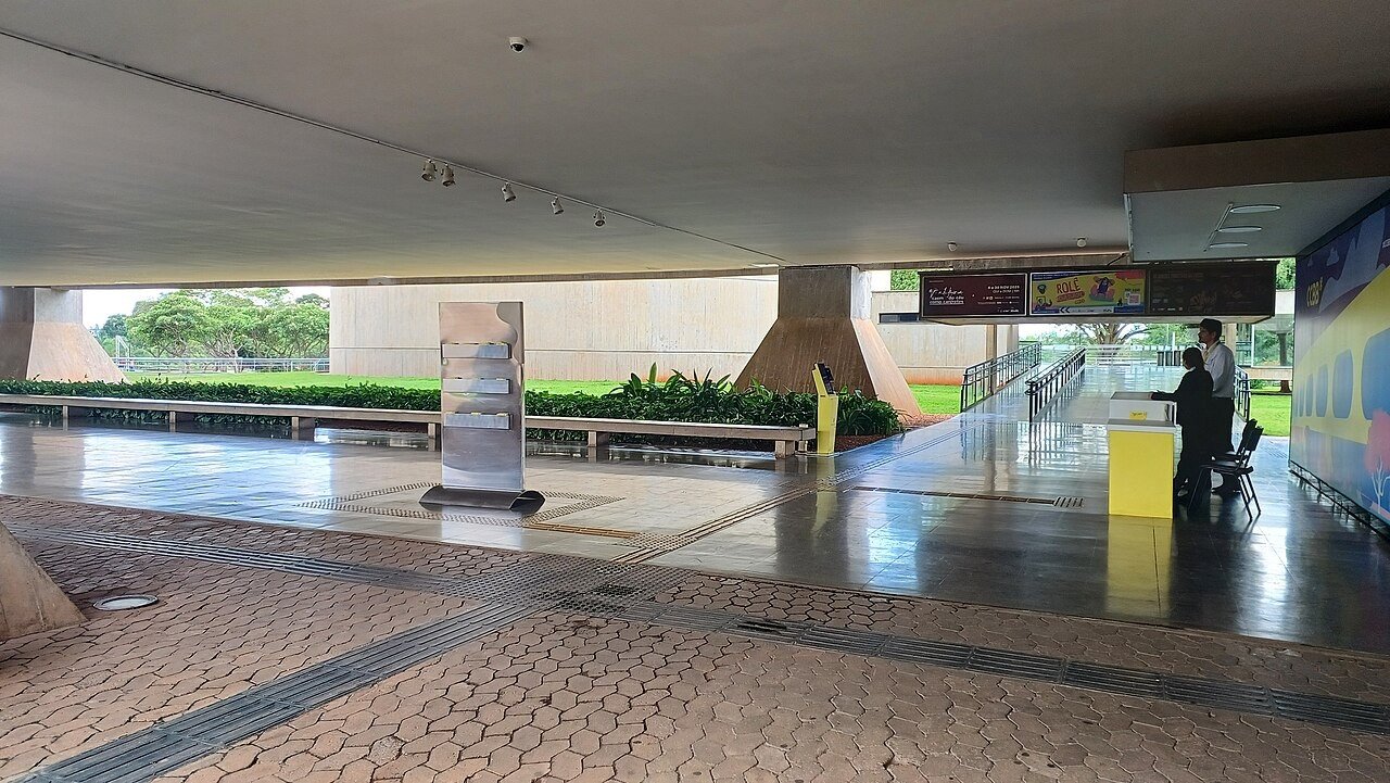 Área coberta de entrada do CCBB Brasília, com colunas de concreto, balcão amarelo de atendimento e cartazes da programação
