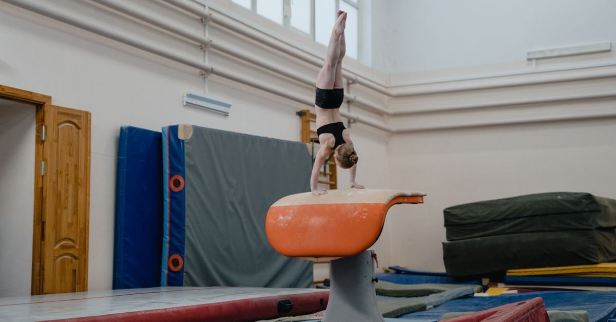 Ginasta faz parada de mão sobre a mesa de salto em ginásio de ginástica artística, com colchões e espaldar ao fundo