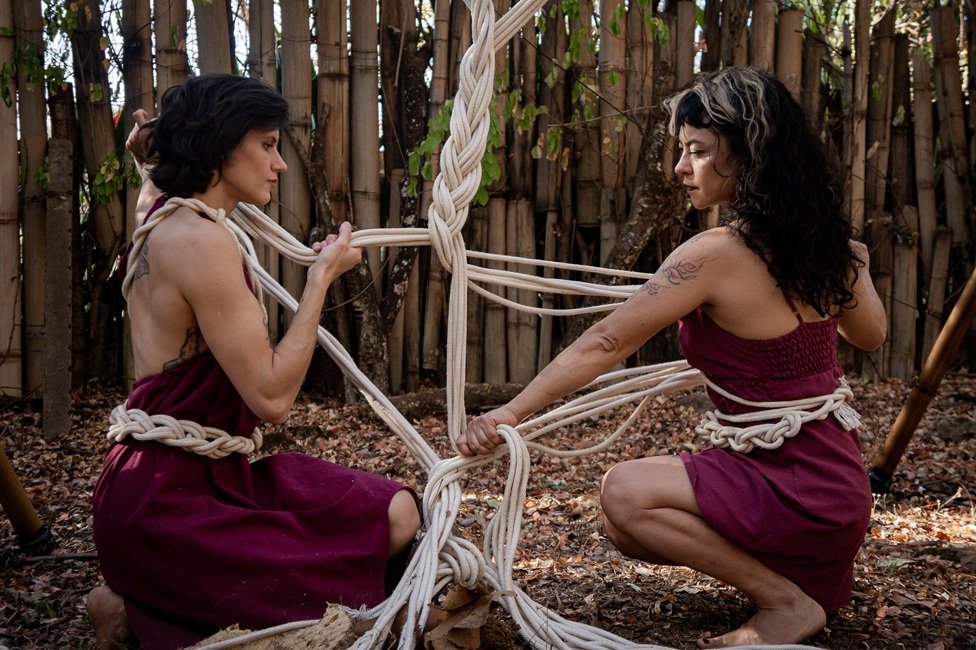 Duas artistas agachadas trançam cordas de macramê entre estruturas de bambu, em area arborizada