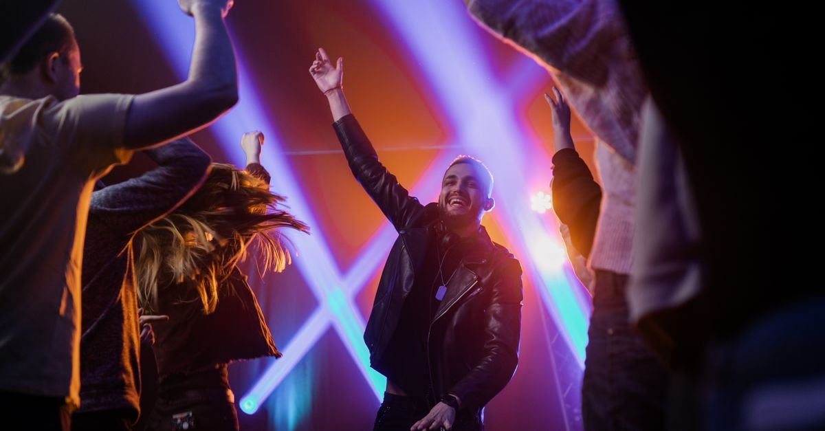 Jovem sorri com a mão levantada em pista de dança iluminada por luzes coloridas