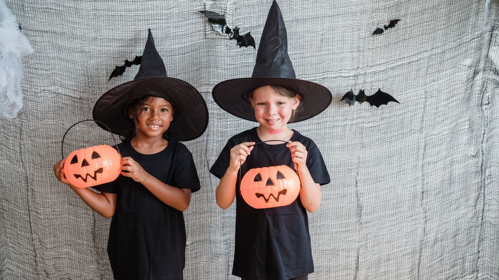 Duas crianças fantasiadas de bruxa seguram abóboras de Halloween diante de um fundo com morcegos de papel