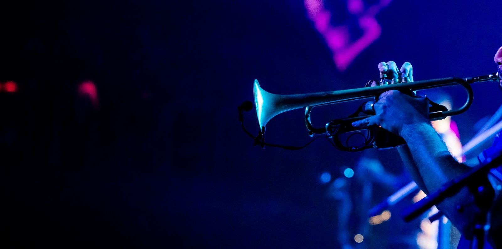 Trompetista toca em palco com luz azul e roxa, em foto de apresentação ao vivo
