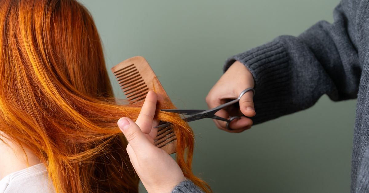 Mãos aparando mecha de cabelo comprido com tesoura e pente de madeira
