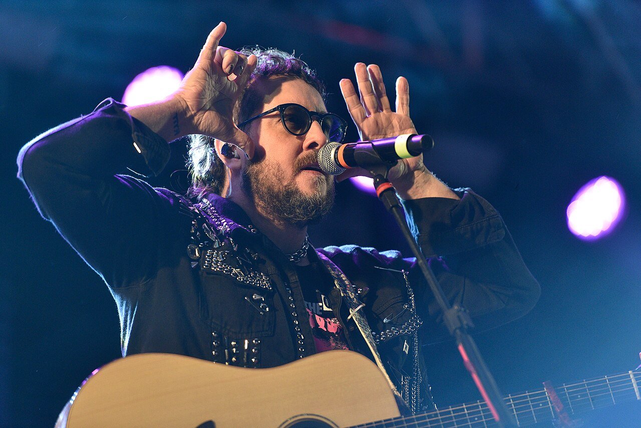 Vocalista do Barão Vermelho canta ao microfone com violão durante show, sob luz azul de palco