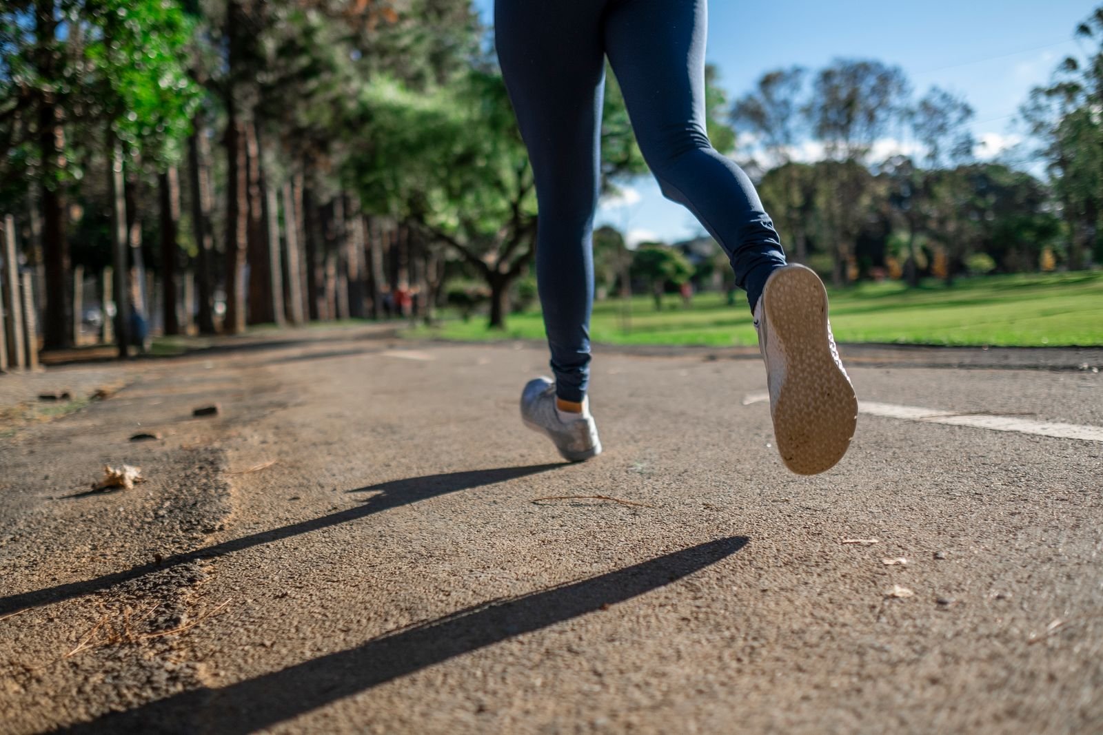 Corredora de legging azul em pista de asfalto cercada de árvores, foto ilustrativa de corrida em parque
