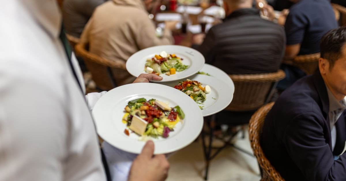 Garçom serve dois pratos de salada a clientes sentados em mesas de restaurante