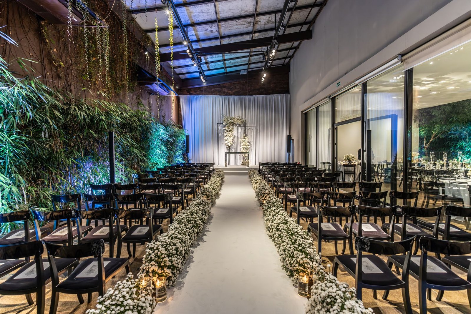 Salão de cerimônia de casamento montado com cadeiras, corredor de flores brancas e altar ao fundo