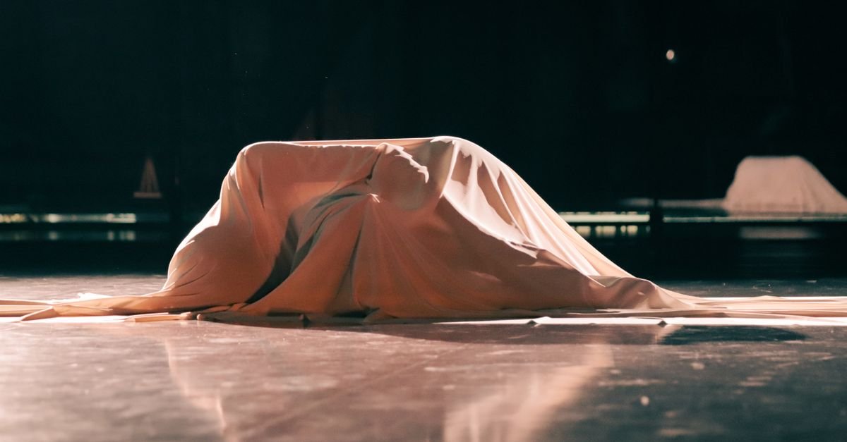 Artista coberta por um tecido claro em ação de performance sobre o palco iluminado