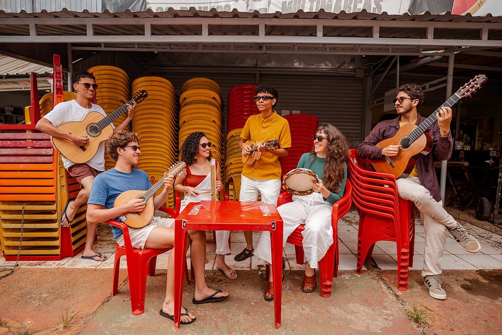 Seis músicos tocam violão, bandolim, cavaquinho e pandeiro sentados em cadeiras de plástico na área de uma feira