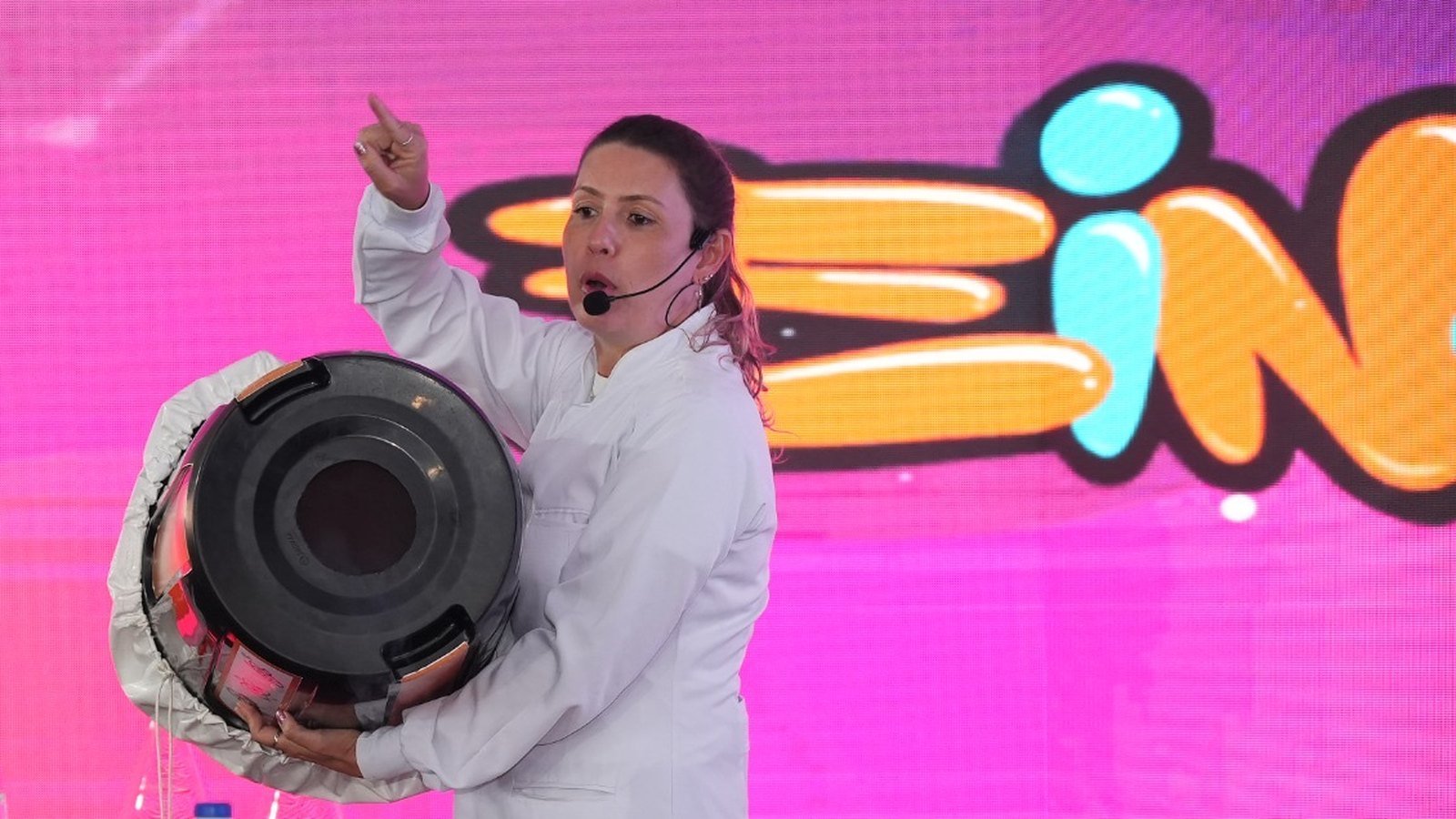 Apresentadora de jaleco branco segura canhão de ar em show de ciência diante de painel com o logo do Einstein Jr.