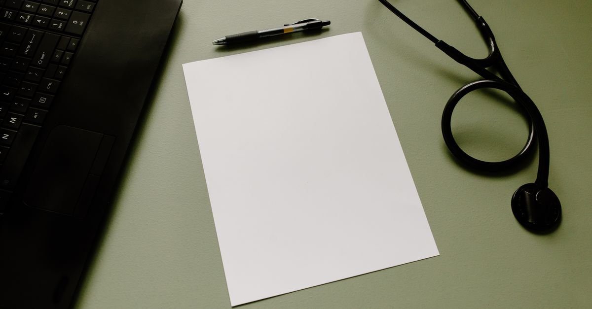 Estetoscópio, caneta e folha de papel em branco sobre mesa de trabalho ao lado de notebook