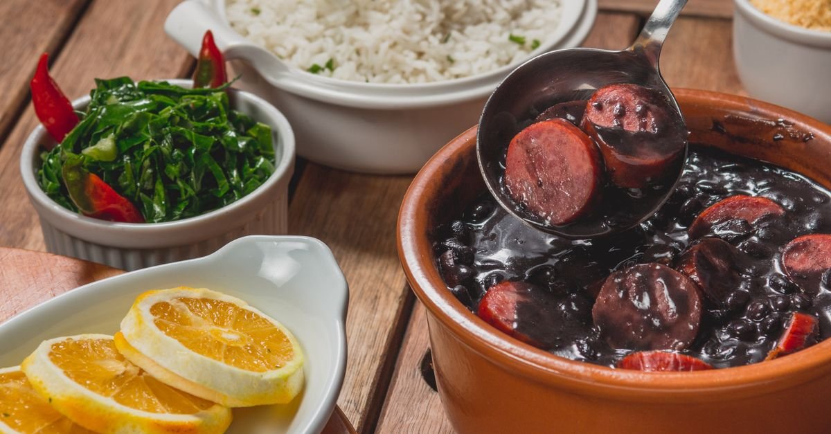 Panela de barro com feijoada, ao lado de arroz, couve refogada e fatias de laranja