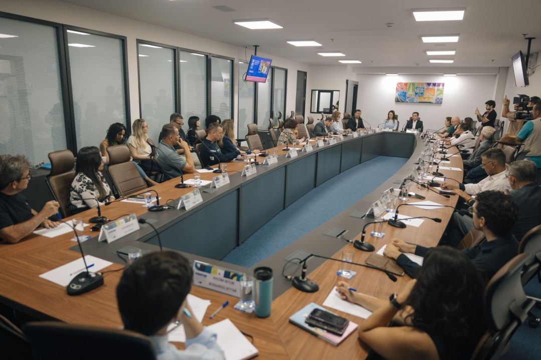 Conselheiros sentados em mesa em U durante a reunião do Condetur, com o secretário de Turismo ao centro