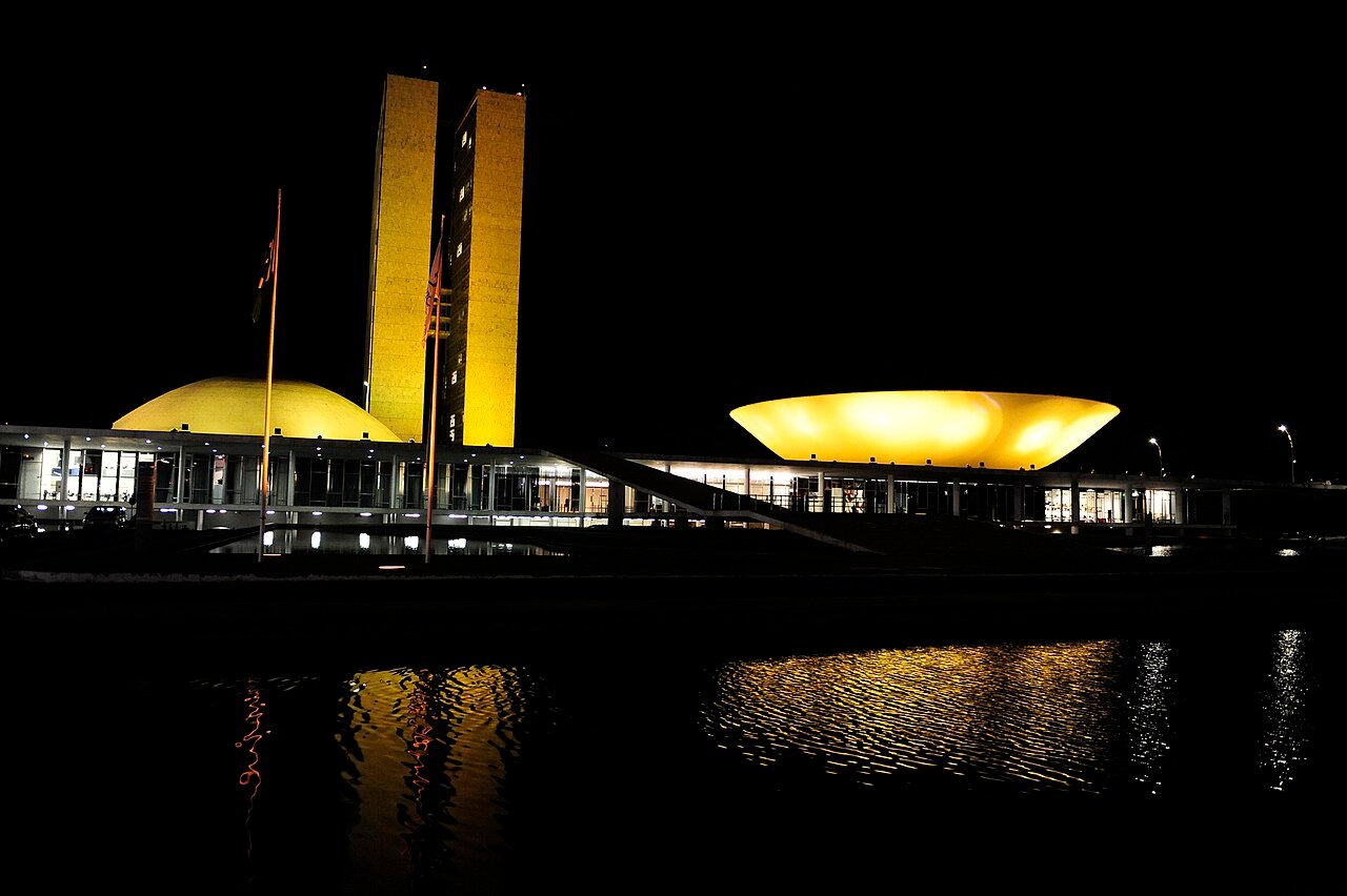 Fachada do Congresso Nacional iluminada de amarelo à noite, com reflexo no espelho d'água