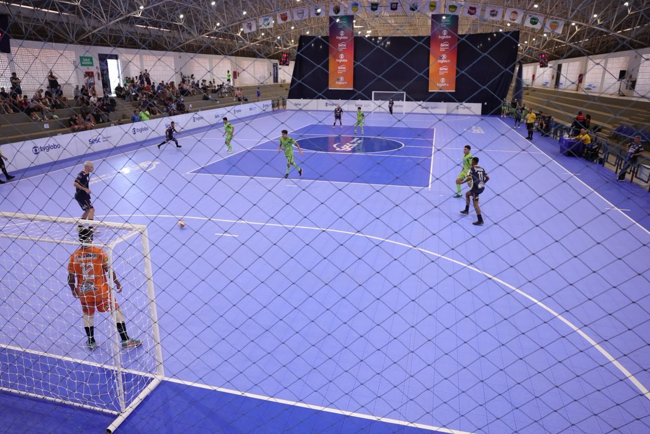 Partida de futsal na quadra azul do Ginásio do Sesc Ceilândia, com bandeirolas das regiões administrativas no teto e arquibancada cheia