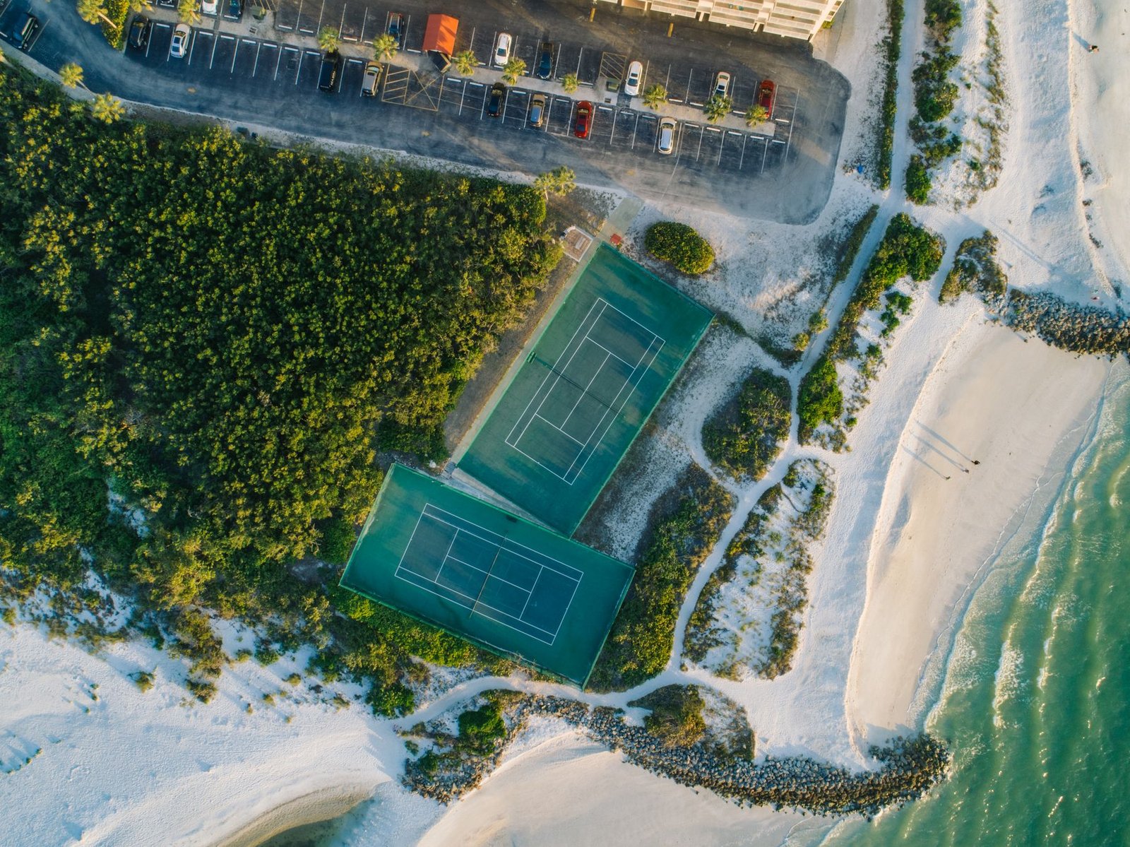 Vista aérea de duas quadras de tênis ao lado de uma praia, imagem ilustrativa do clima de esporte de praia