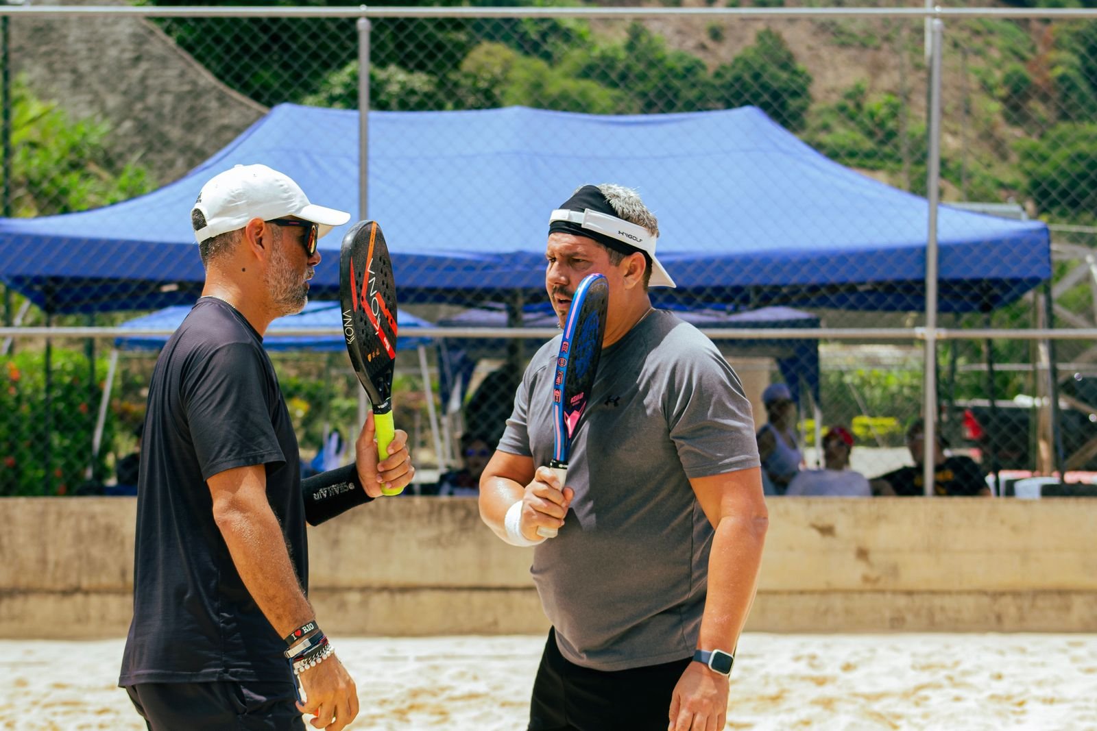 Dois jogadores de beach tennis seguram raquetes em quadra de areia, em dia de sol