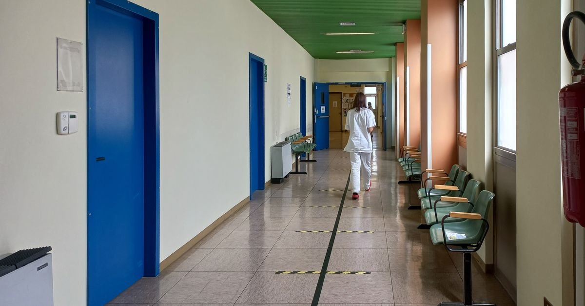 Profissional de saúde de uniforme branco caminha por corredor de hospital com portas azuis