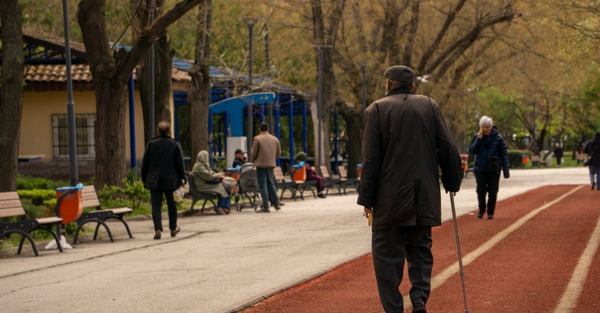 Homem idoso caminha com bengala ao lado de uma pista vermelha de corrida em parque arborizado