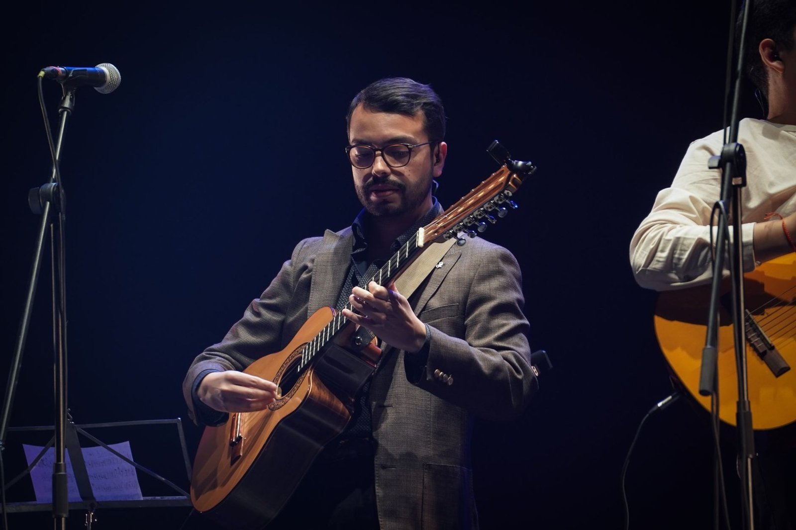 Músico de óculos toca violão em palco escuro, ao lado de um microfone