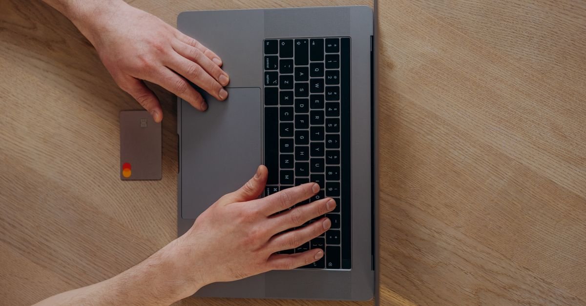 Vista de cima de mãos sobre o teclado de um notebook ao lado de um cartão bancário