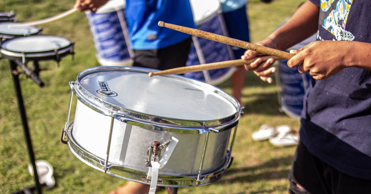 Mãos tocando caixa de bateria com baquetas durante ensaio de percussão ao ar livre