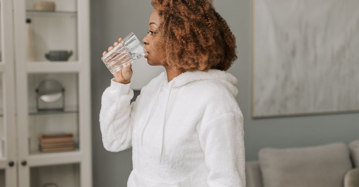 Mulher de moletom branco bebe agua em copo de vidro dentro de casa
