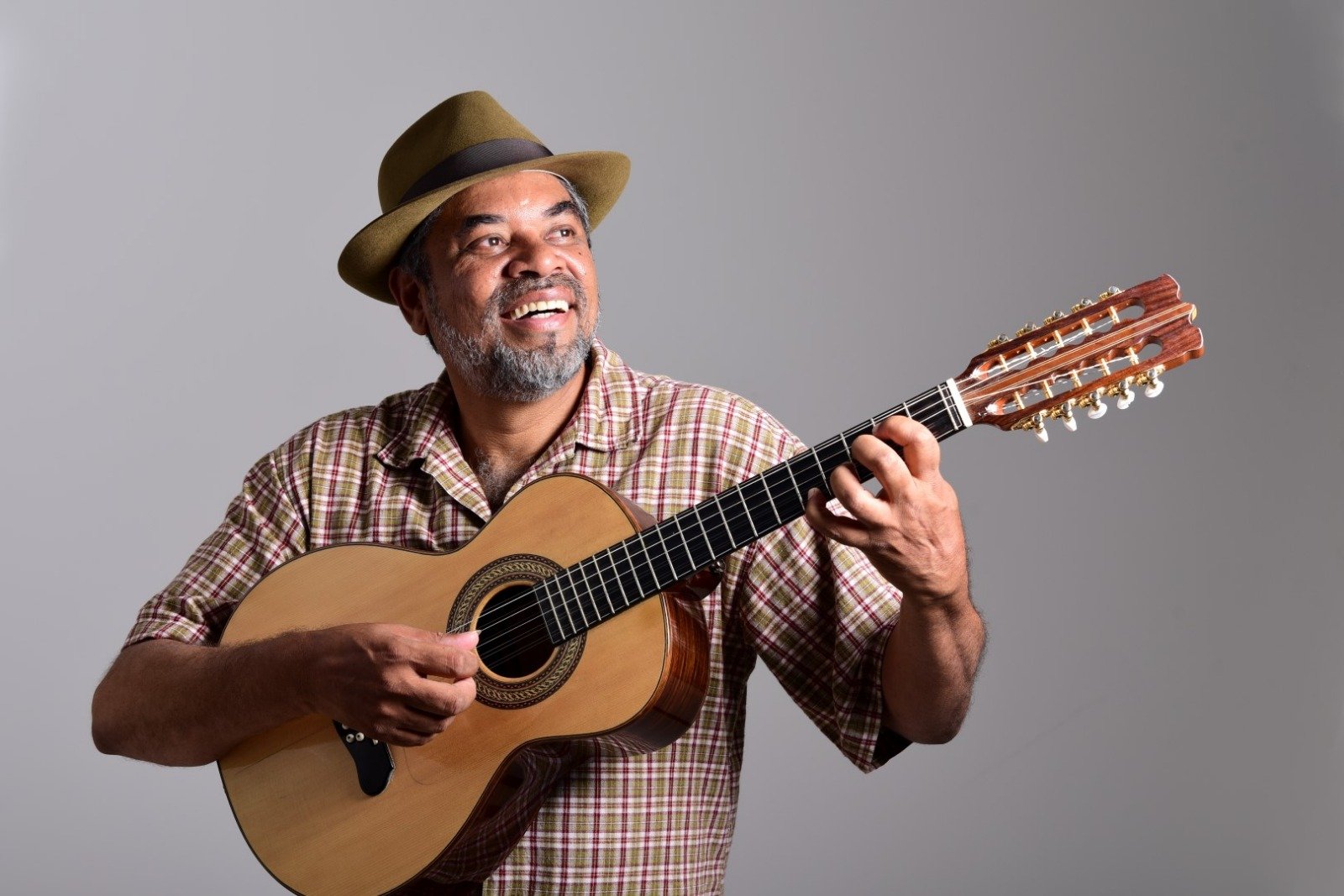 O musico Volmi Batista, de chapeu e camisa xadrez, toca viola de dez cordas sorrindo, em fundo cinza