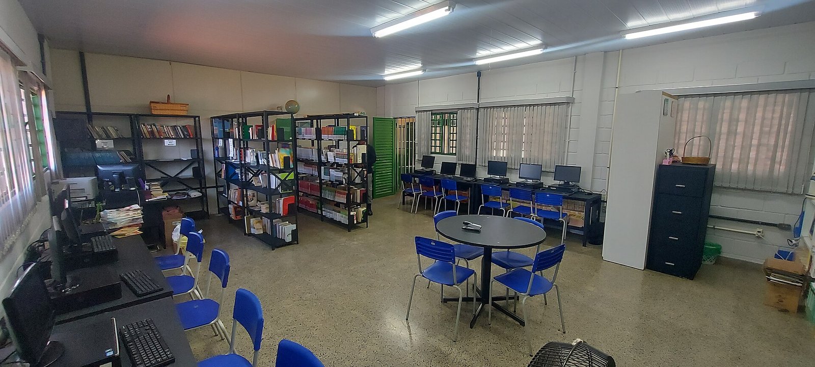 Sala de leitura e informática de escola pública do Distrito Federal, com estantes de livros, computadores e mesa redonda com cadeiras azuis