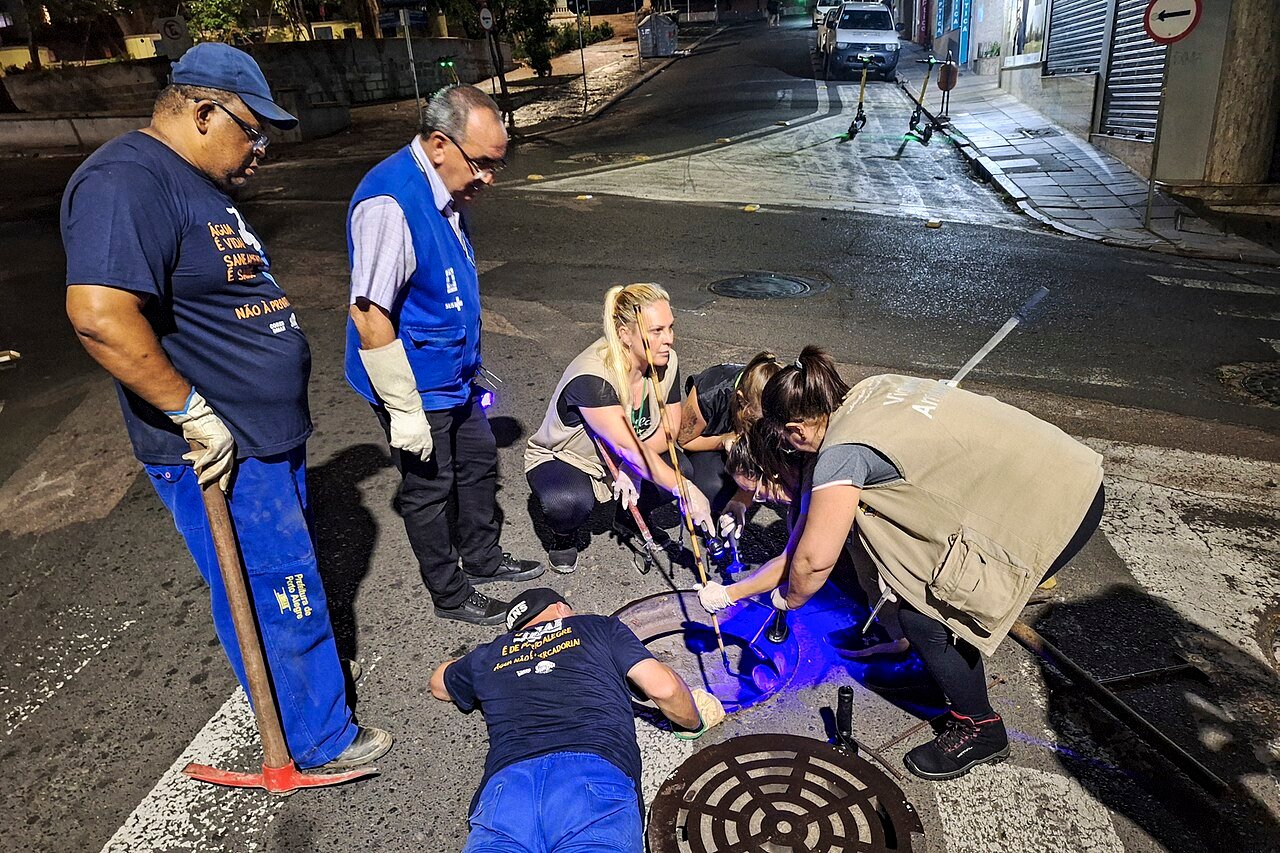 Equipe de vigilância ambiental abre uma caixa de esgoto na rua à noite, com lanterna de luz ultravioleta