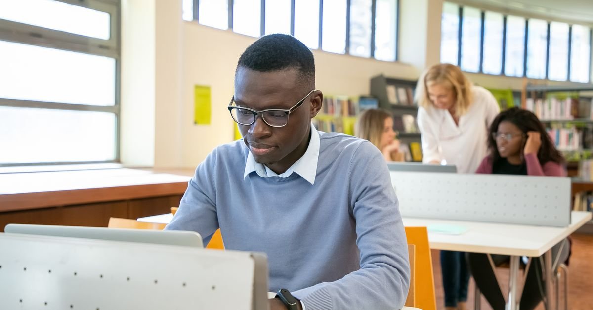 Estudante de óculos usa notebook em mesa de biblioteca, com outros estudantes ao fundo