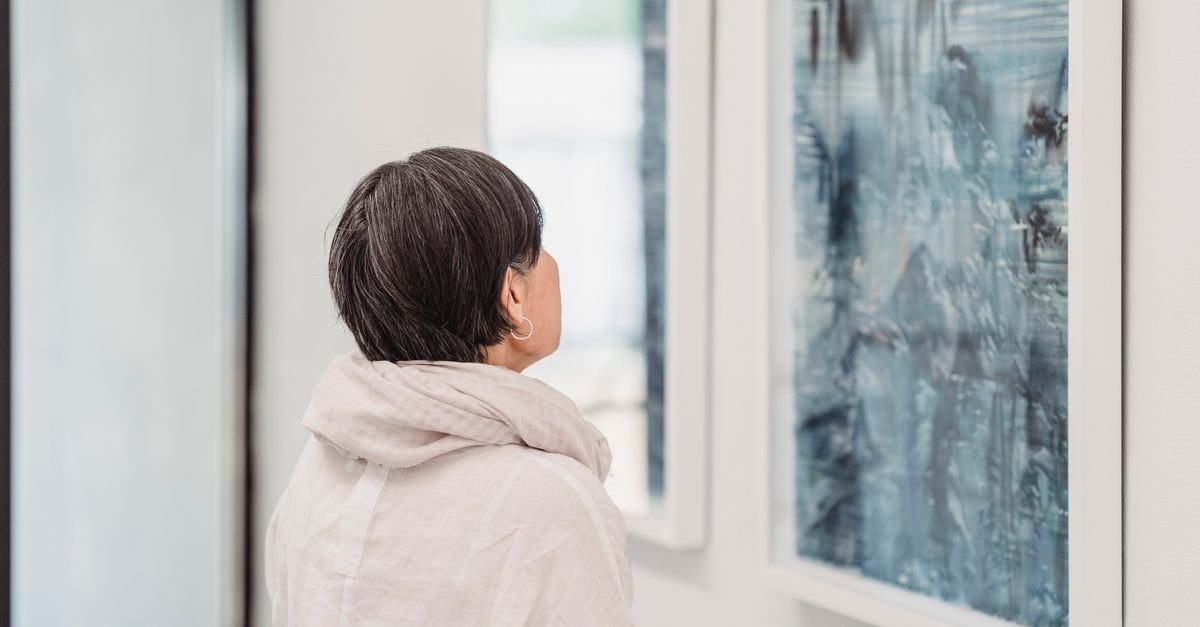Mulher observa pintura abstrata em tons de azul emoldurada na parede branca de uma galeria de arte
