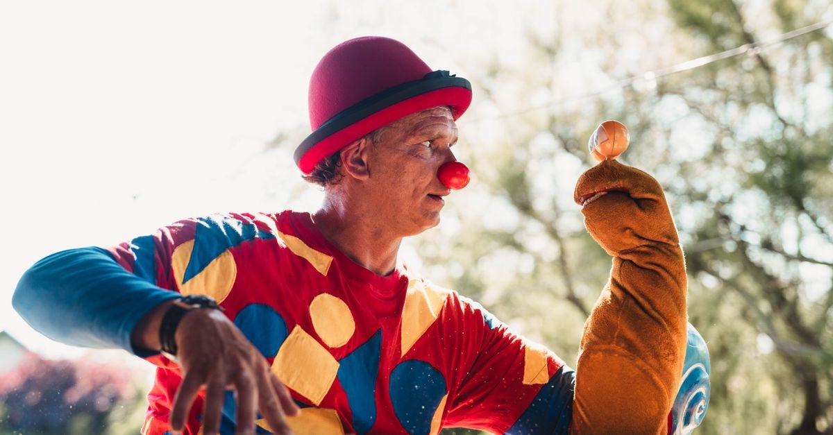 Palhaço com nariz vermelho e chapéu-coco apresenta número com fantoche ao ar livre