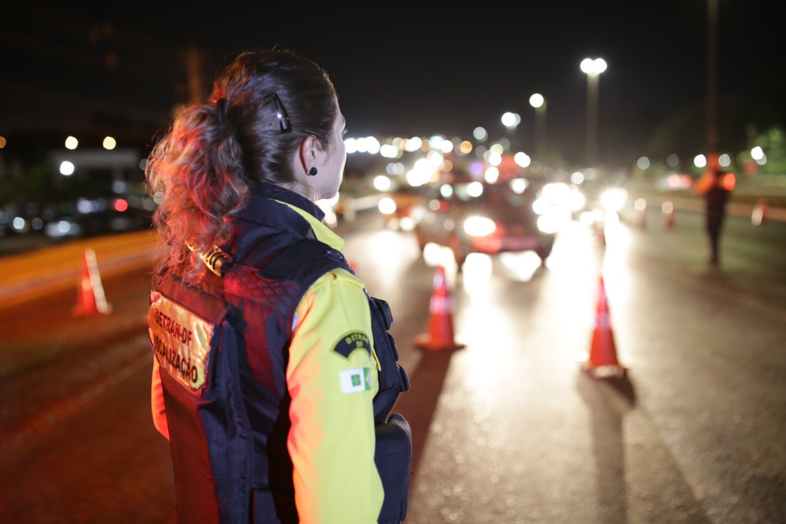 Agente do Detran-DF de costas, com colete de fiscalização, observa a pista durante blitz noturna com cones e faróis de veículos
