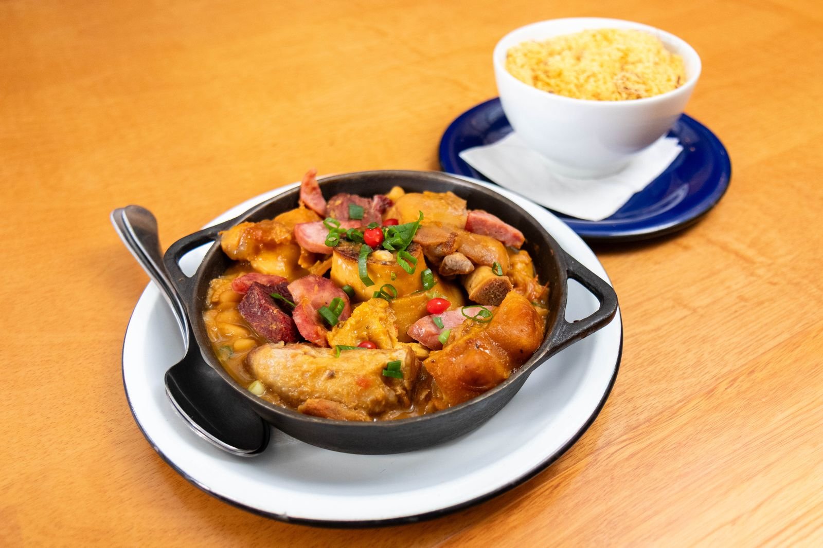 Feijoada servida em panela de ferro preta, com farofa em uma tigela ao fundo (foto ilustrativa)