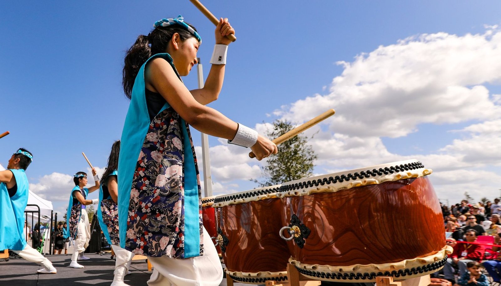 Grupo de taiko toca tambores japoneses em palco ao ar livre durante festival cultural, com plateia sentada ao lado