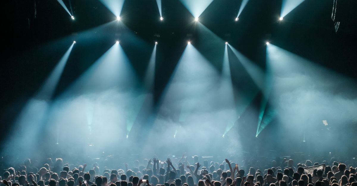 Público de costas diante de palco iluminado por refletores azuis e fumaça durante show noturno