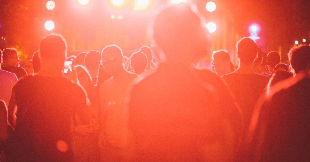 Público de costas diante de palco iluminado em festival ao ar livre à noite