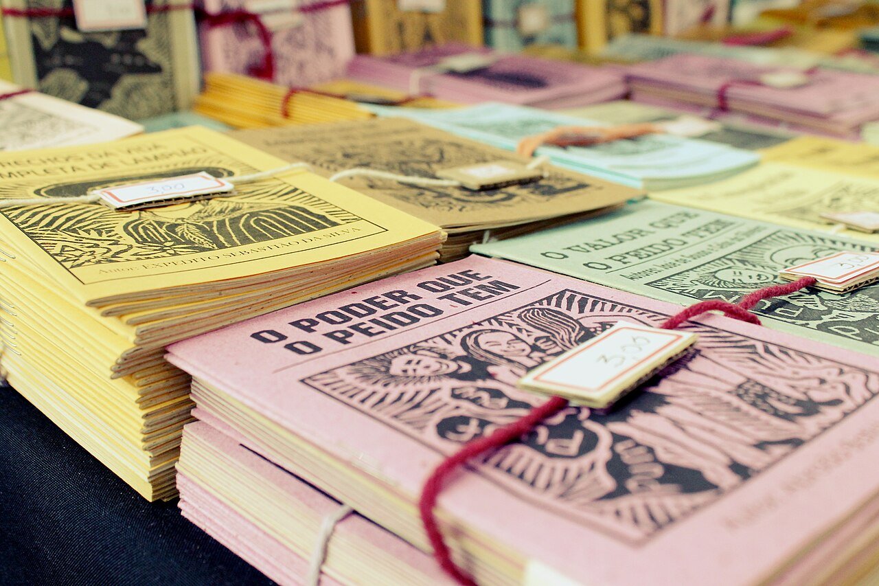 Folhetos de literatura de cordel empilhados sobre mesa, com capas de xilogravura e etiquetas de preço