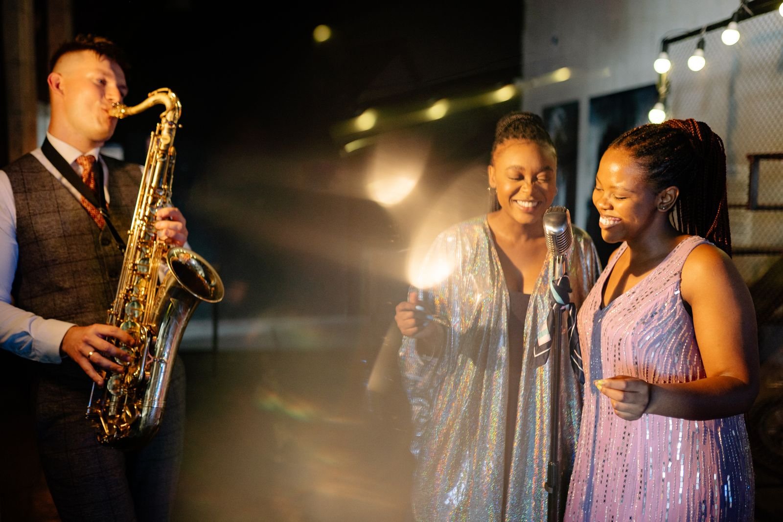 Saxofonista e duas cantoras sorrindo diante de um microfone de pedestal em um palco de casa de shows (foto ilustrativa)
