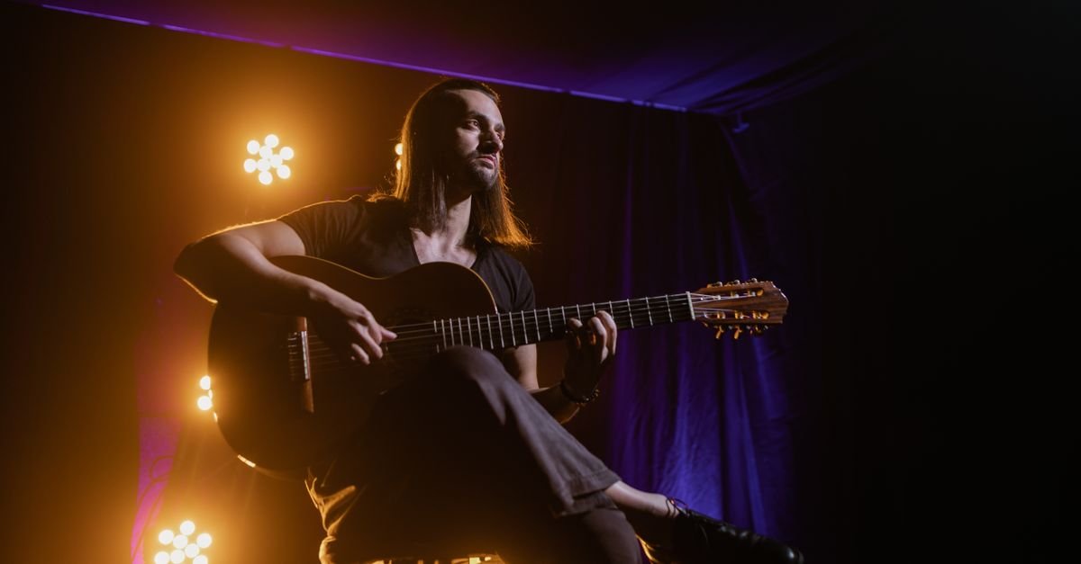 Músico toca violão de nylon sentado em palco iluminado por luzes laranja e roxa