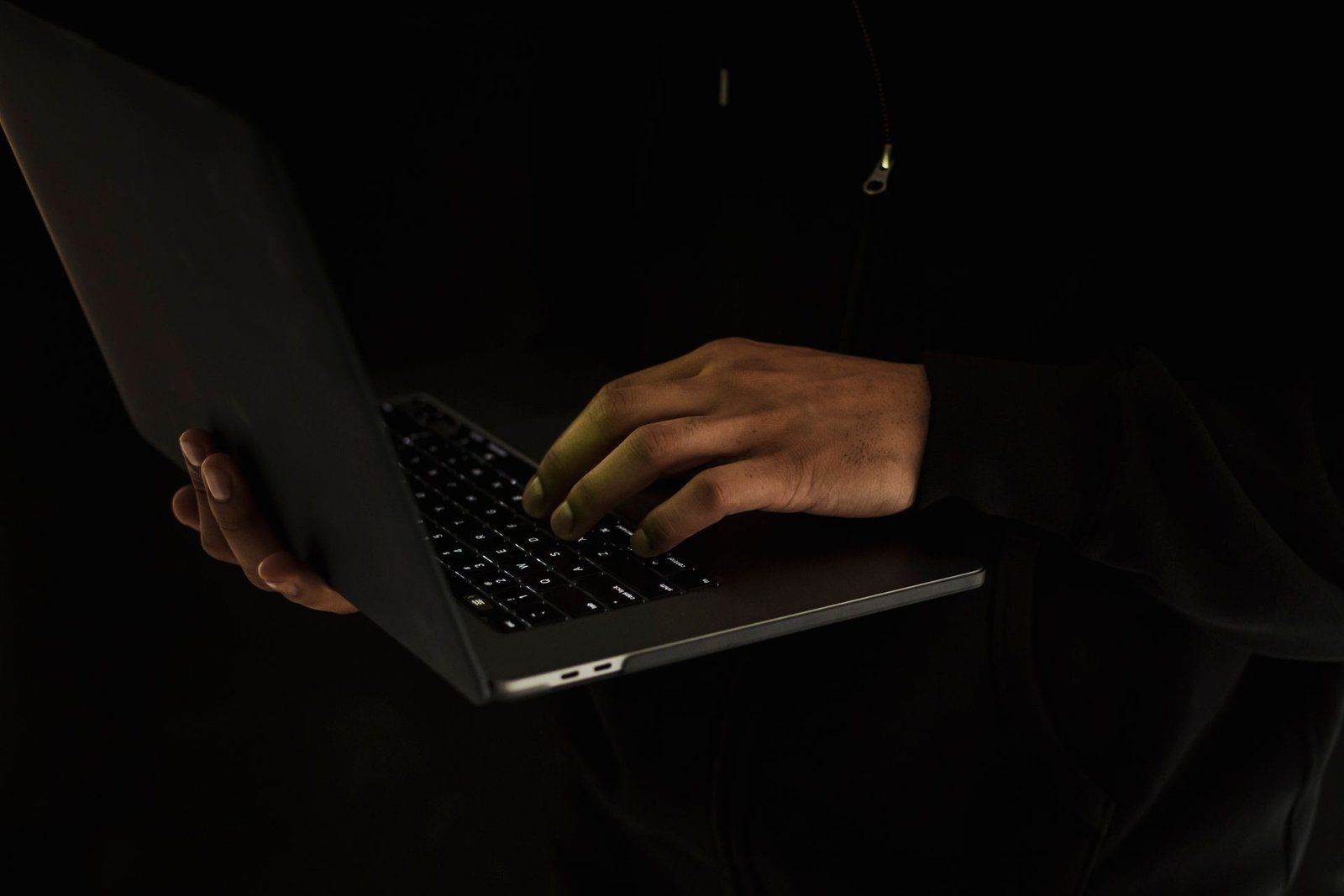 Mãos de uma pessoa não identificada digitando em um notebook, em ambiente escuro