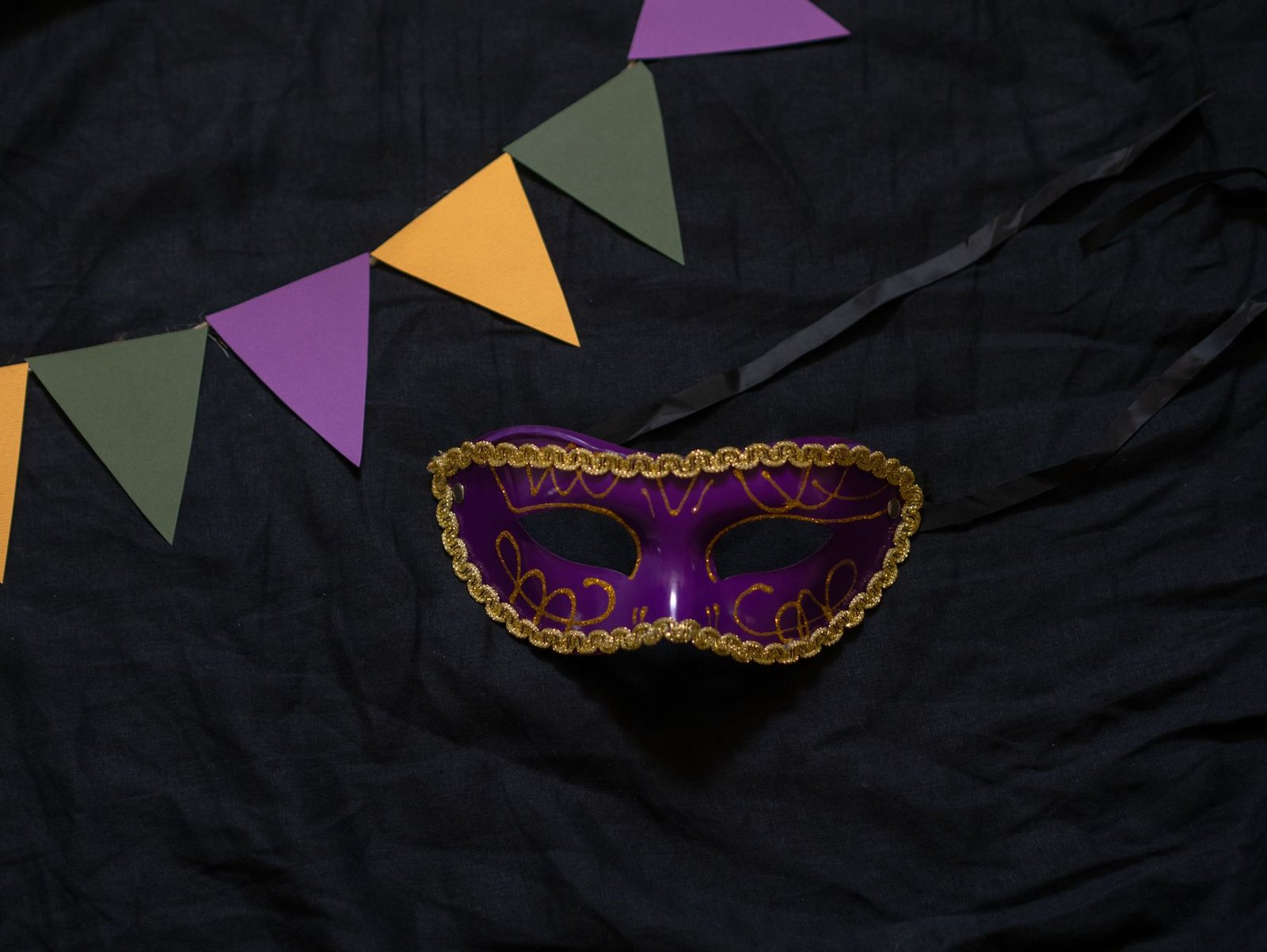 Máscara de baile roxa com acabamento dourado sobre tecido preto, ao lado de bandeirolas de papel