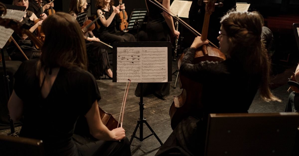 Músicos de orquestra tocando instrumentos de corda diante de partitura em estante durante concerto