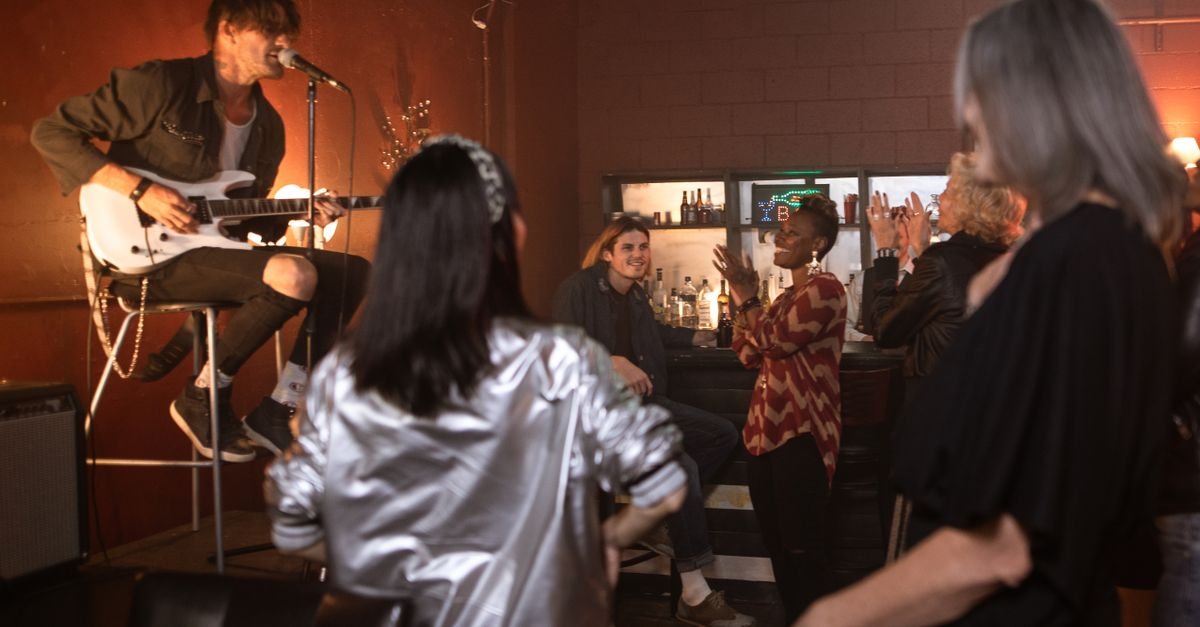 Músico toca guitarra e canta em palco de bar pequeno enquanto o público acompanha de pé